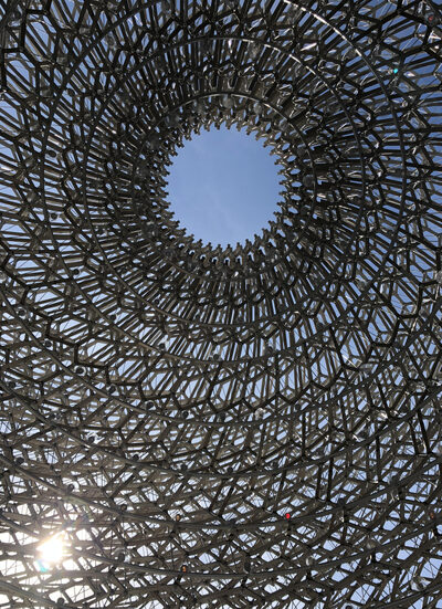 Close-up view of an intricate metallic lattice structure with a circular opening at the top, revealing a clear blue sky and sunlight streaming through the geometric framework